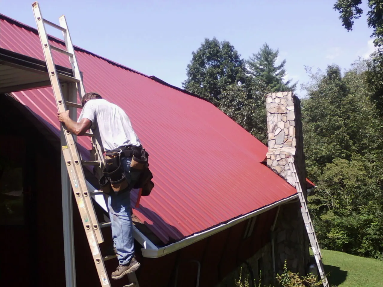 Roofer on a ladder beside a red metal roof and stone chimney — Soffit & Fascia Repair by Roofing Brothers in Jenkinsburg